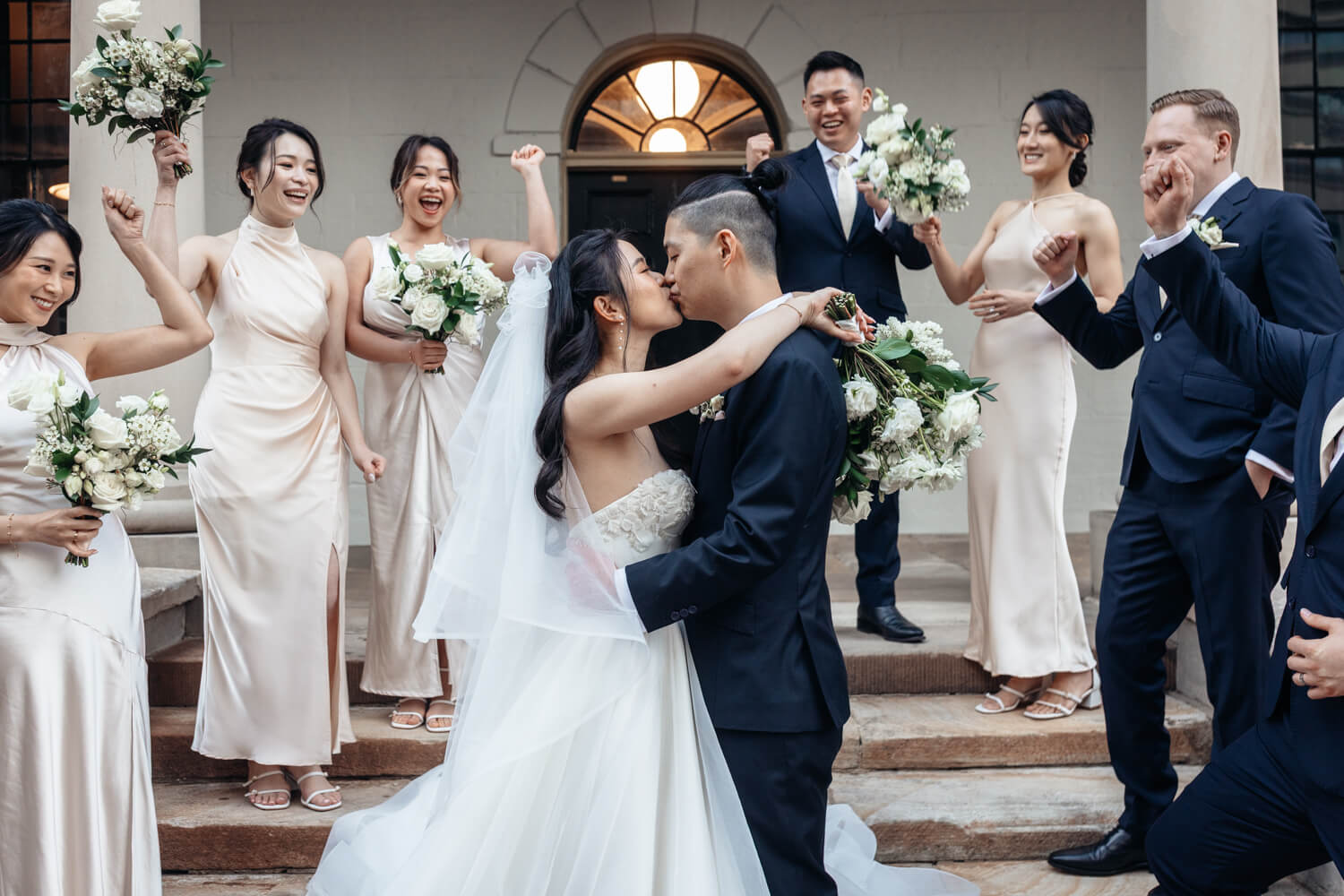 A picture at the Mint of the bride and groom kissing with their bridal party cheering them on